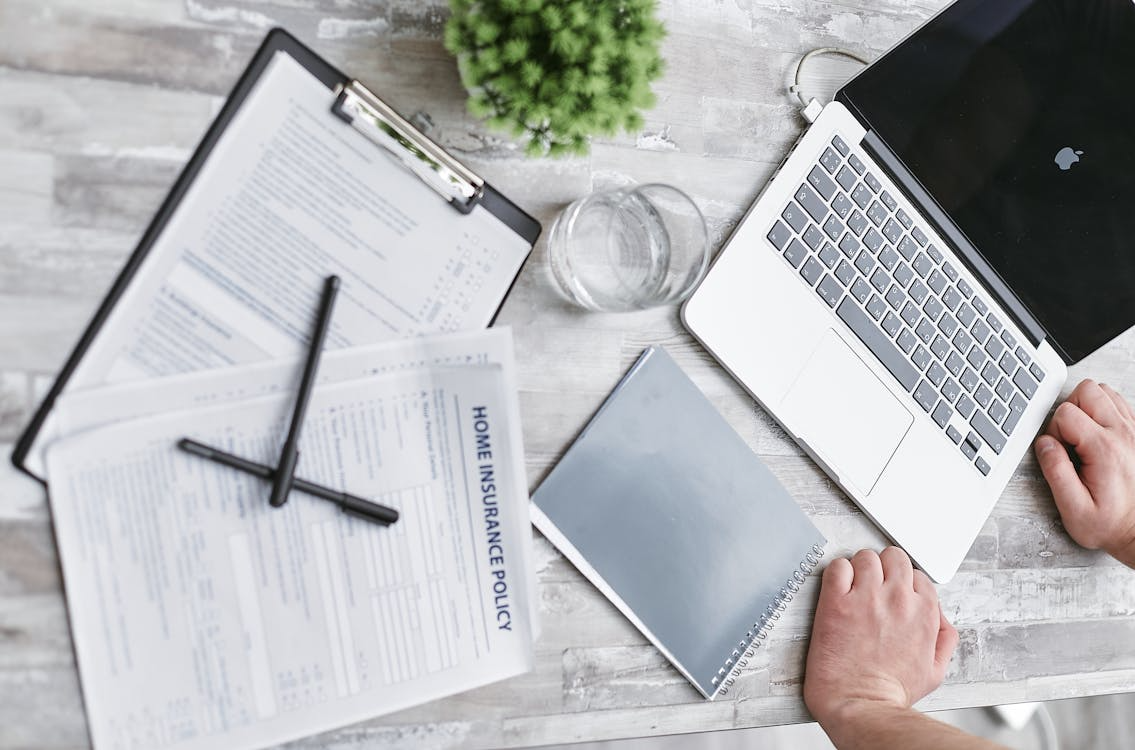 person writing on white paper beside clear glass mug and silver macbook