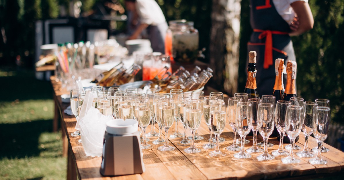 A bartender stands behind an outdoor mobile bar. The bar is a wooden table with champagne glasses, buckets of beer, and more.