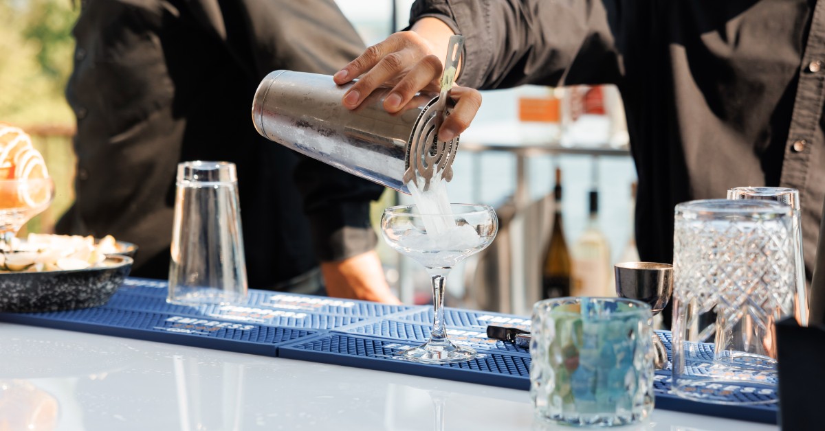 A close-up of a bartender in a black shirt at an outdoor mobile bar pouring a clear cocktail from a strainer into a glass.
