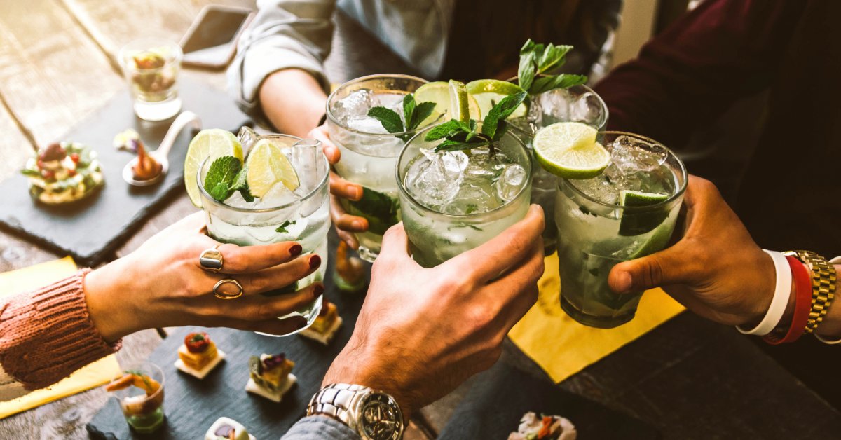 An overhead view of a group of people toasting each other over a table with cocktails featuring mint and lime garnishes.