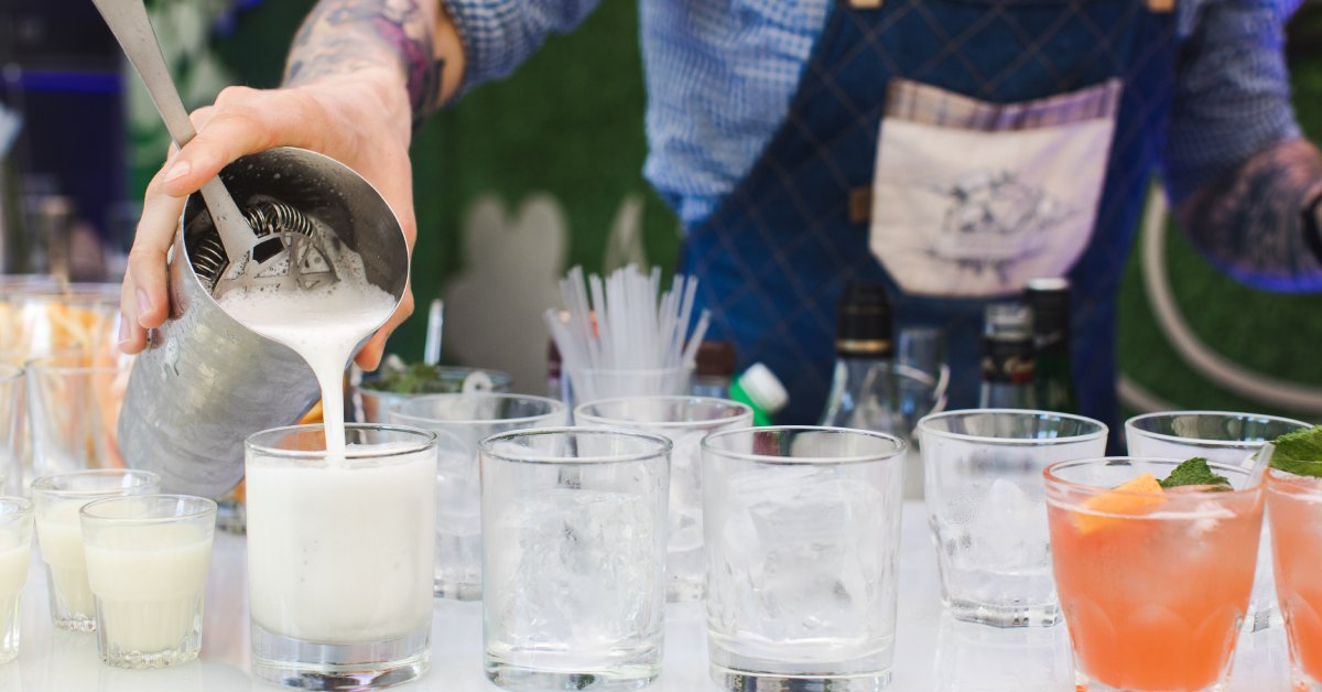 A close-up of a bartender's hand as he pours a cocktail from a mixer and strainers into a glass on a table full of drinks.