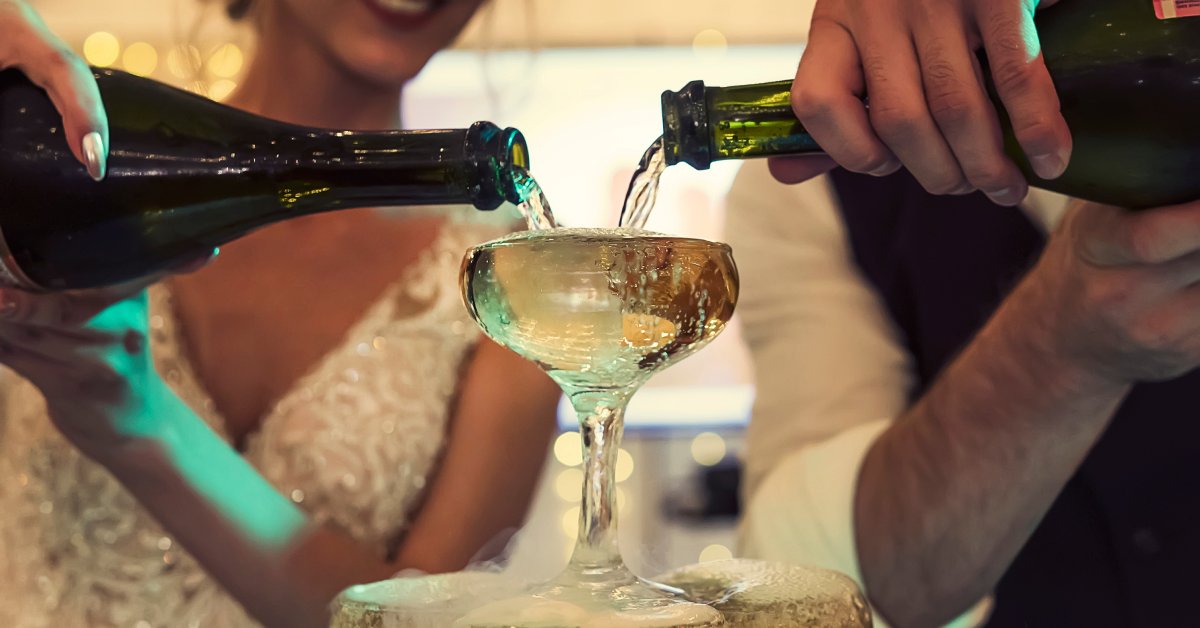 A close-up of the top glass of a champagne tower as the bride and groom pour from bottles into the glass.