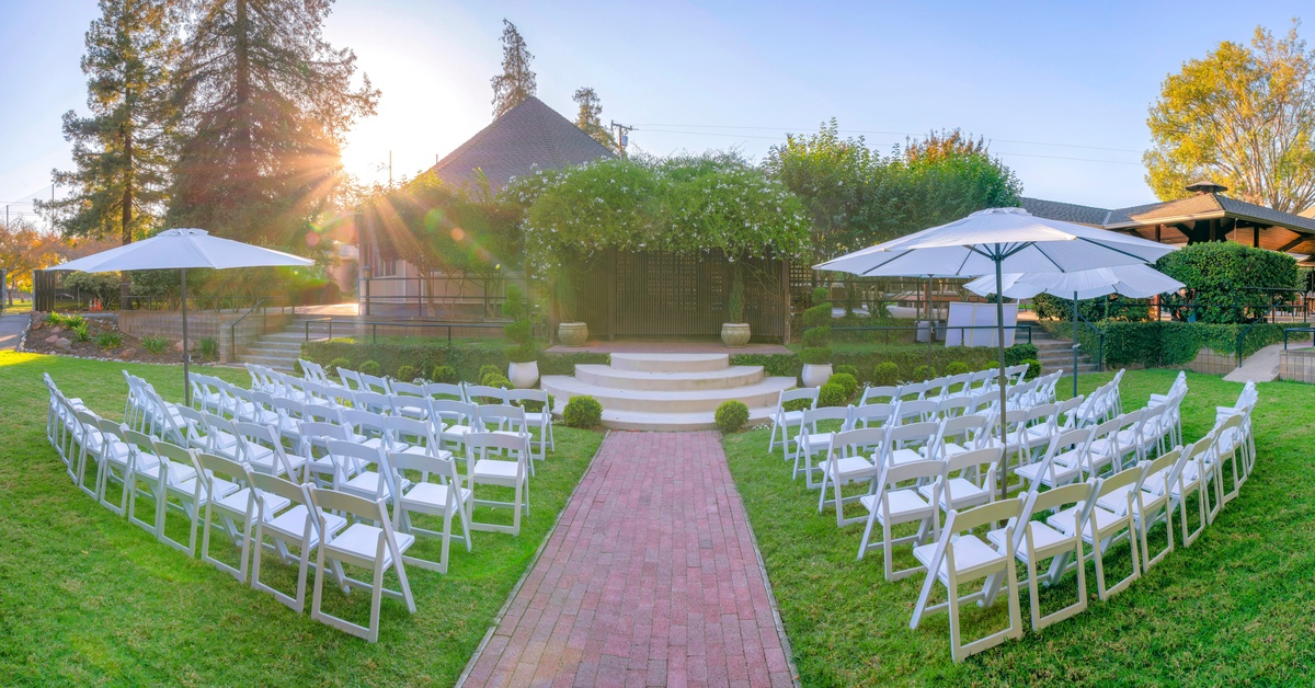 A view of a garden wedding venue from the center brick aisle with white chairs on either side and umbrellas.