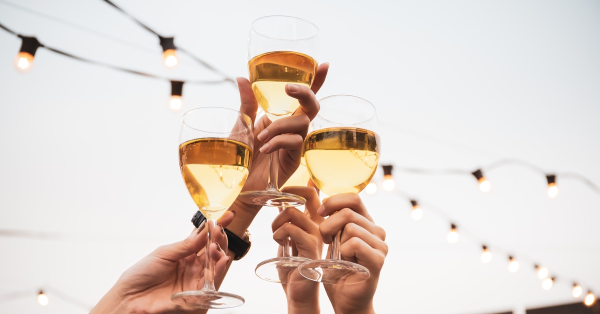 A close-up of four people holding up their wine glasses together in a toast with a white tent and festival lights behind.