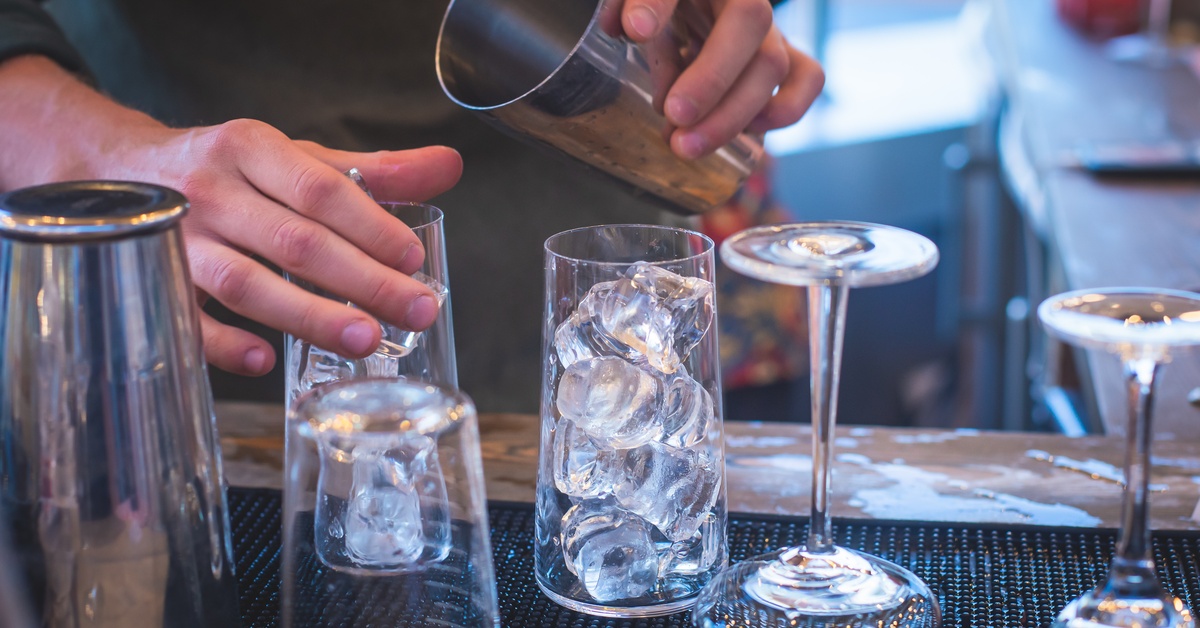 A close-up of a bartender's hands as they make a cocktail. They are in the middle of pouring a drink into a glass of ice.