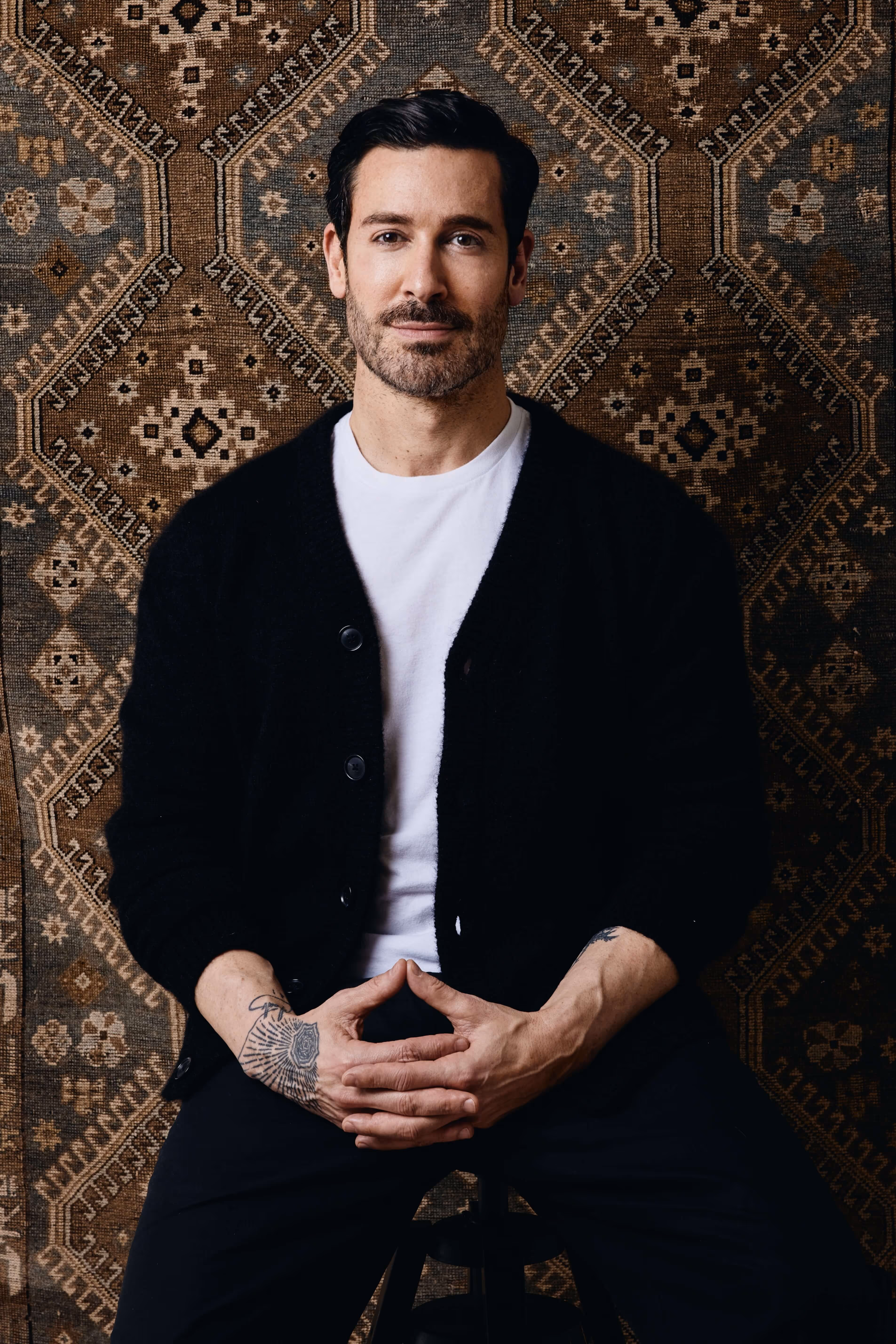 Jason Moattar portrait in front of vintage rug backdrop, wearing a light knit sweater and showcasing wrist tattoo; rug designer and textile expert.