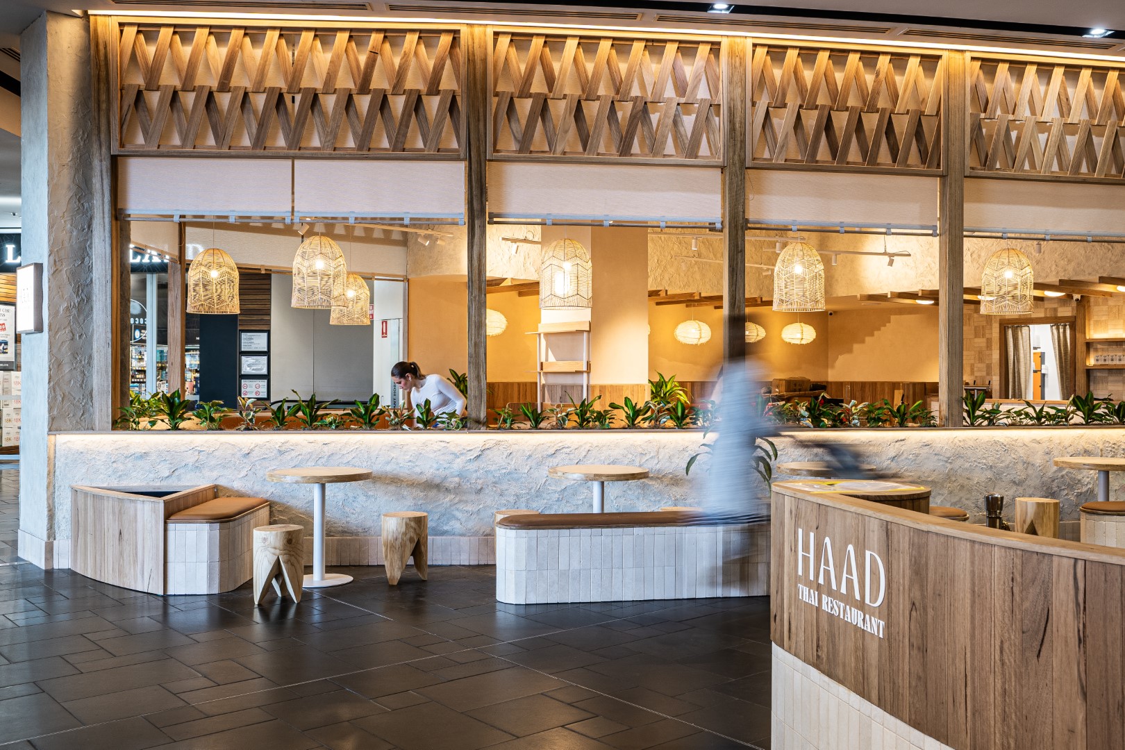 Interior of Haad Thai Restaurant with wooden furniture, hanging woven lamps, and a person working behind the counter.