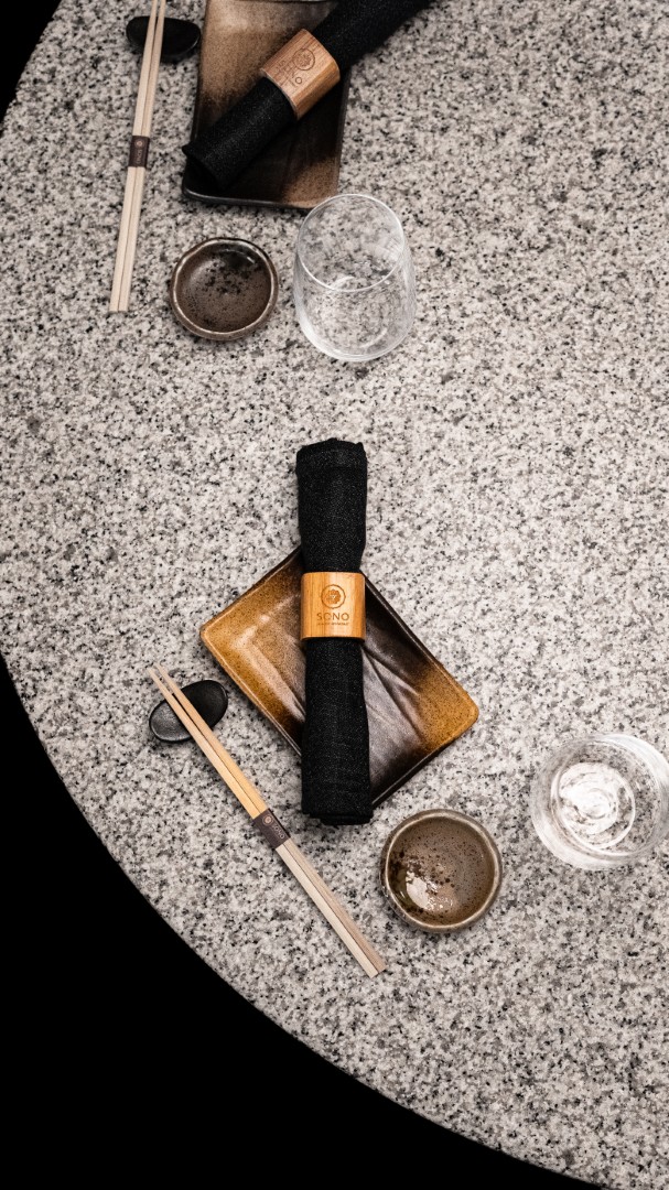 Top-down view of a Japanese table setting with chopsticks, black napkins wrapped in wooden rings on rectangular plates, empty glasses, and small ceramic bowls on a speckled gray stone surface.