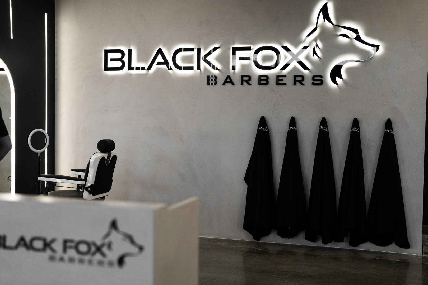 Interior of Black Fox Barbers shop with an empty barber chair, black capes hanging on hooks, and illuminated logo on the wall.