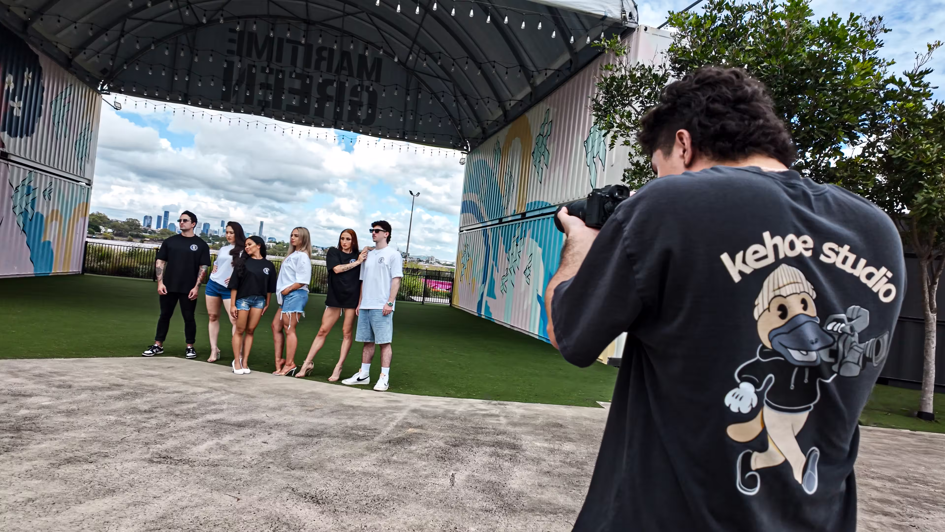 Photographer wearing a 'kehoe studio' shirt takes a photo of six models standing under a covered outdoor area with murals and city skyline in the background.