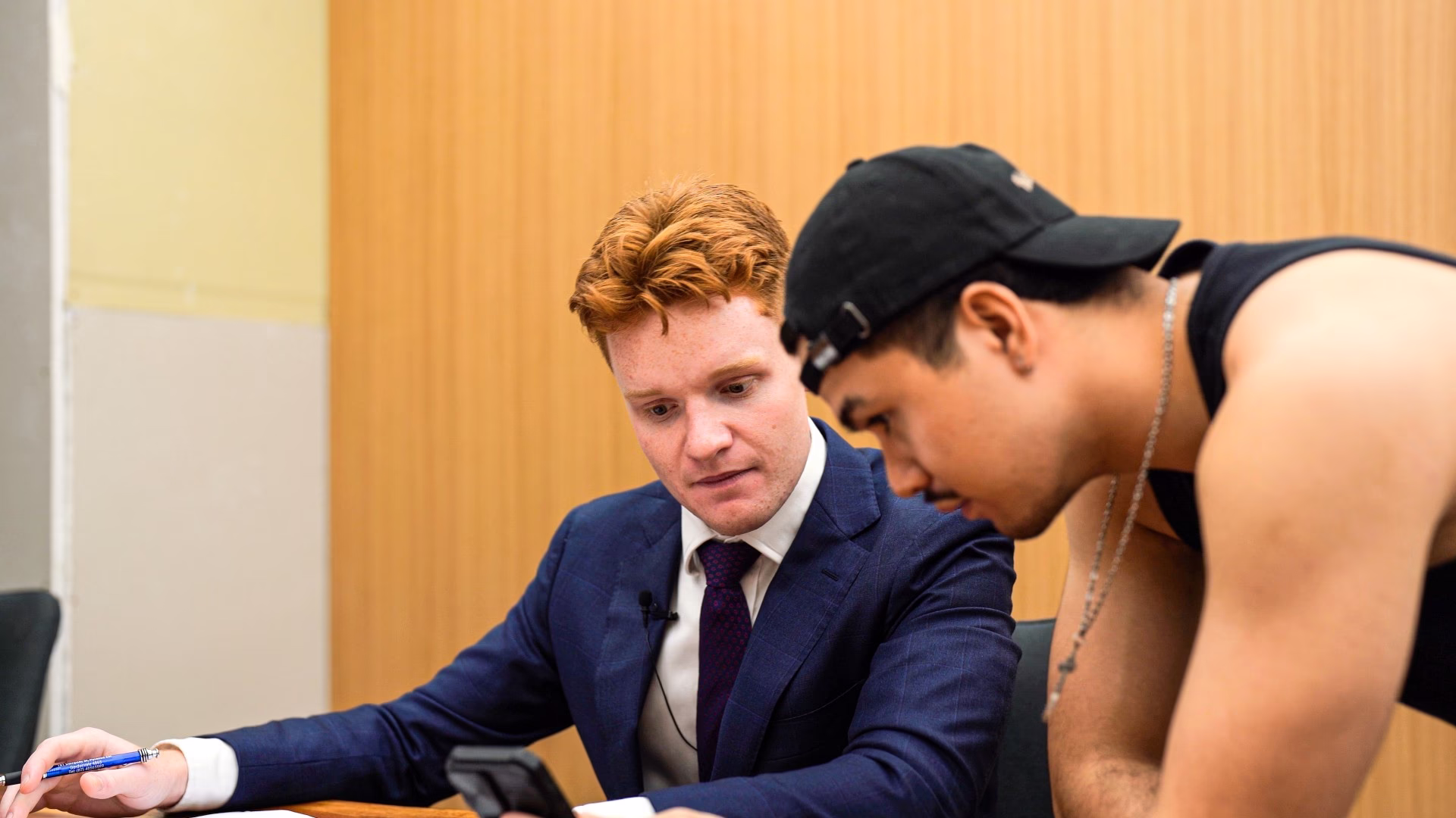 Two men focused on a smartphone, one in a suit and the other in a black tank top and cap.