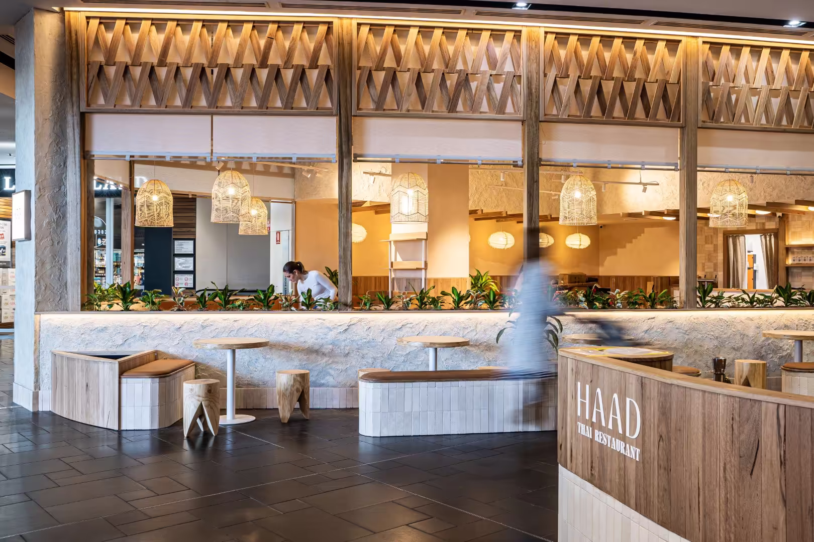 Modern Thai restaurant interior with wooden furniture, hanging woven lamps, plants along a half wall, and a staff member behind the counter.
