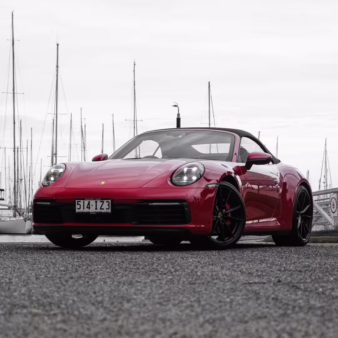 Red Porsche convertible sports car parked near marina with sailboat masts in background.