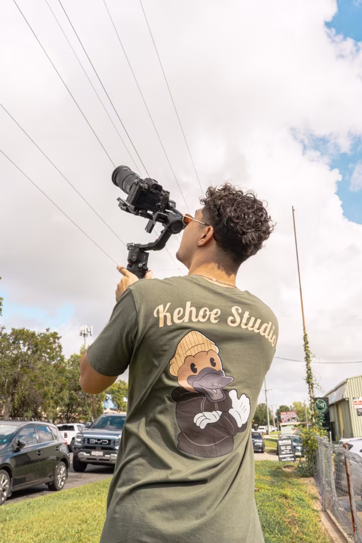 Man with curly hair filming with camera gimbal outdoors wearing green Kehoe Studio shirt with duck logo.