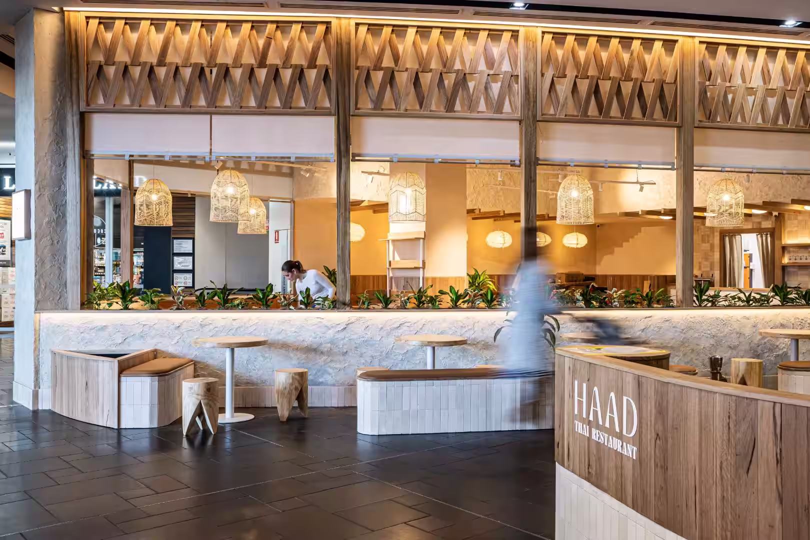 Modern Thai restaurant interior with wooden seating, hanging lamps, plants, and a server behind counter.