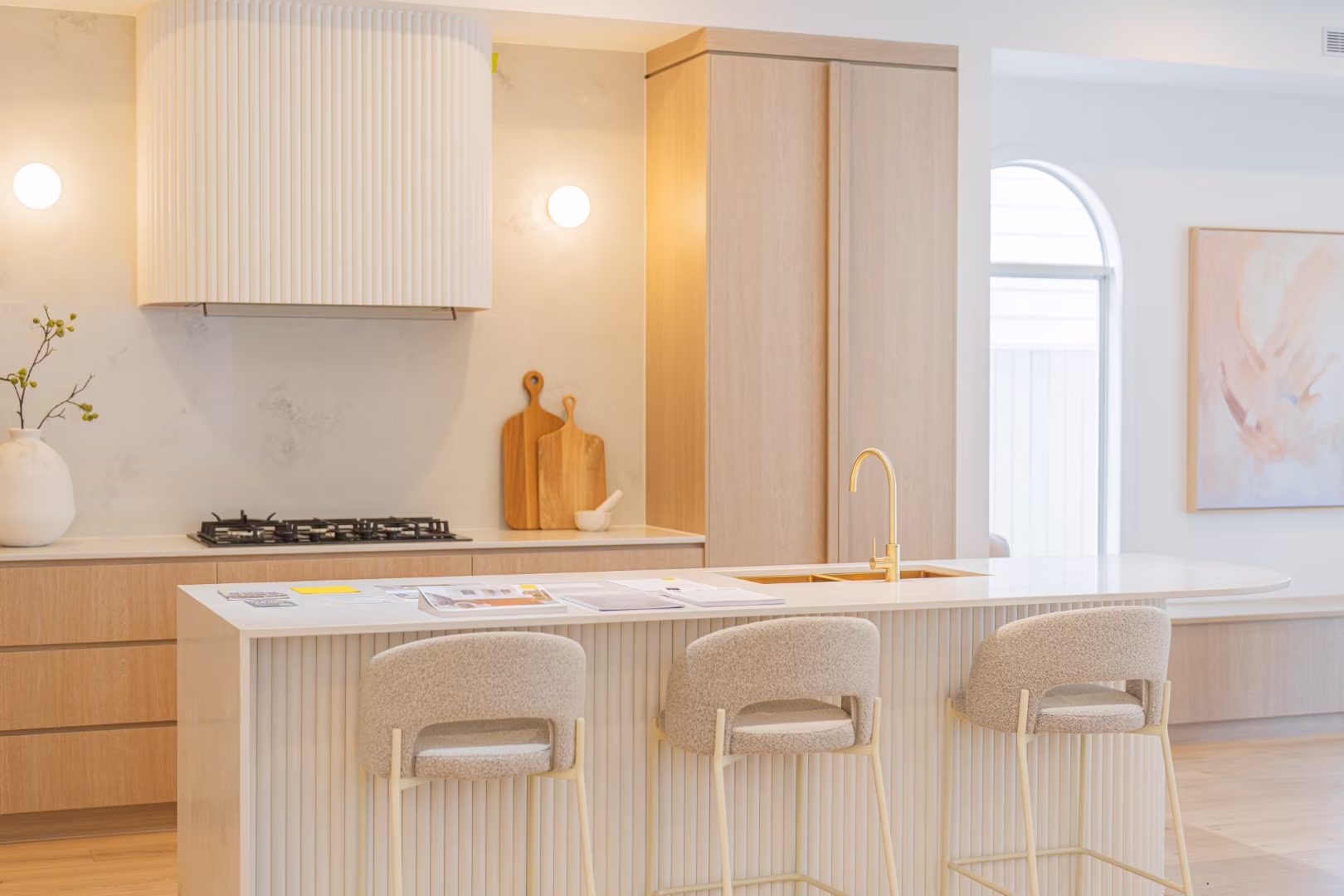 Modern kitchen with light wood cabinets, white island, gold faucet, and three beige bar stools.