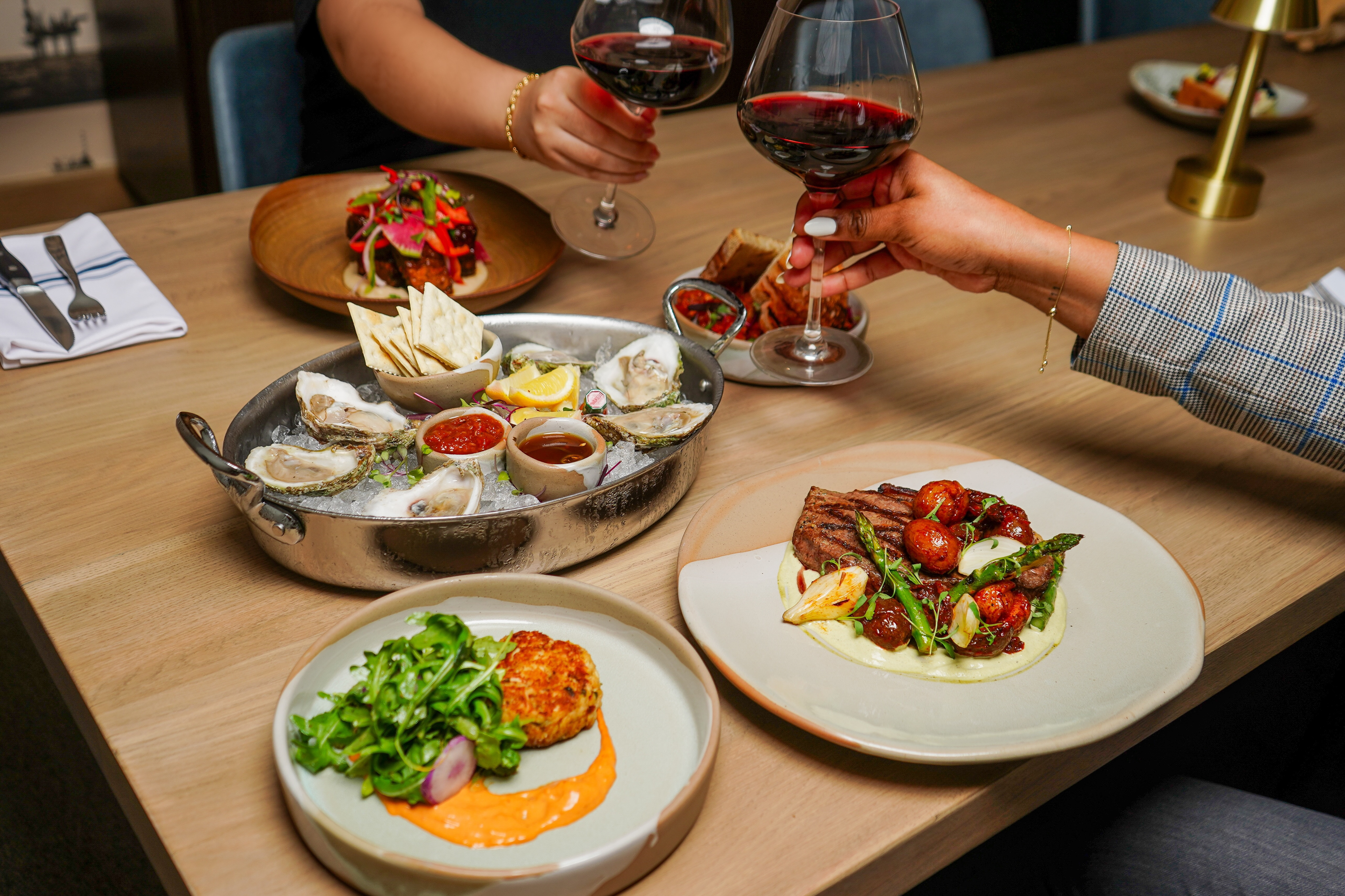 Two guests toasting red wine over oysters, crab cake and steak at a community dinner
