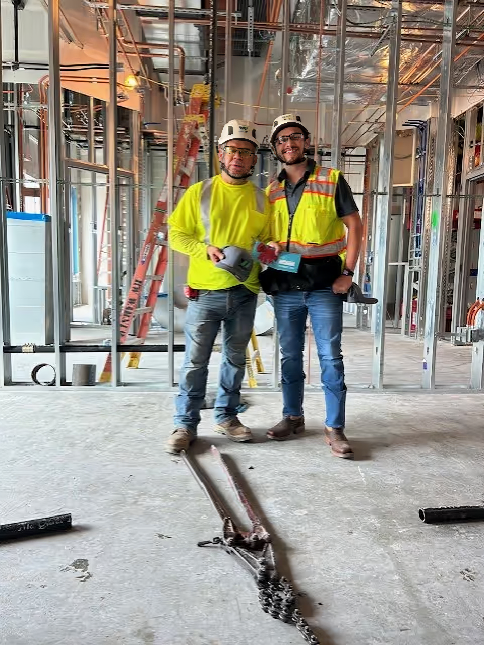 Two construction workers smiling inside a building under renovation, wearing safety gear and standing on a concrete floor.

