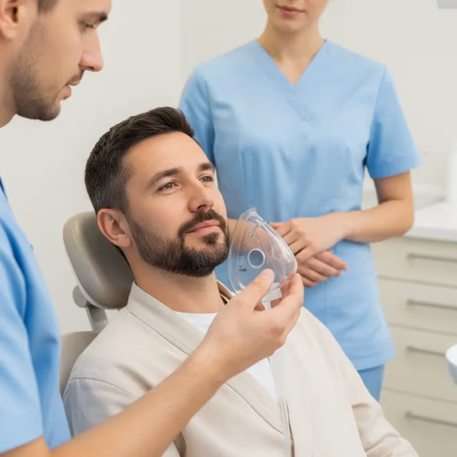 Moški pacient v zobozdravniškem stolu prejema splošno anestezijo skozi masko, obkrožata ga zdravstvena delavca v modrih uniformah.