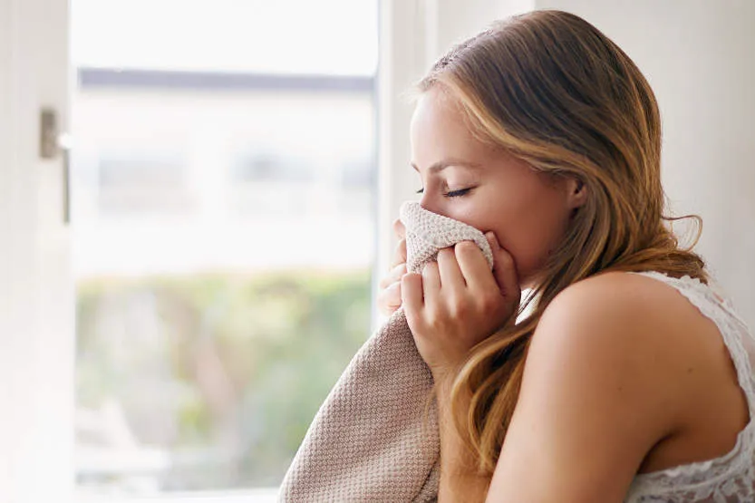 Ženska z dolgimi lasmi diha na mehko tkanino pred odprtim oknom.