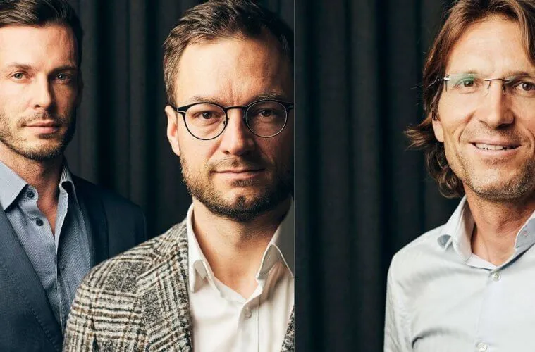 Tri moški v poslovni obleki stoje ob črnem ozadju, eden z očali in brado, drugi z okvirjem in srajco, vsi gledajo v kamero.