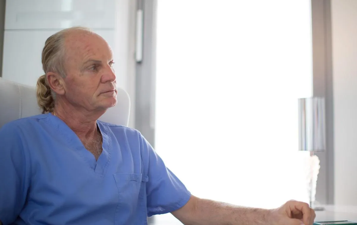 Moški zdravnik v modri medicinski uniformi sedi in zamišljeno gleda v stran.