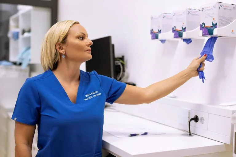 Zdrava ženska v modrih medicinskih oblačilih, ki zajema modro medicinsko rokavico iz stene v bolnišničnem okolju.