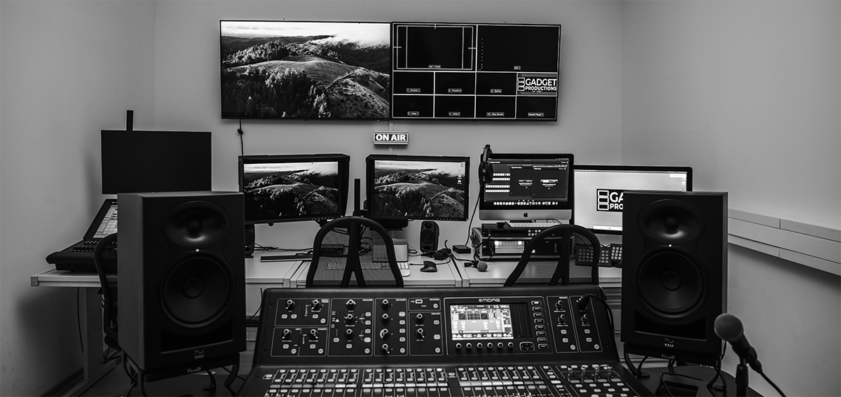 Schwarz-weißes Tonstudio mit Mischpult, zwei großen Lautsprechern, mehreren Monitoren und 'On Air'-Schild an der Wand.