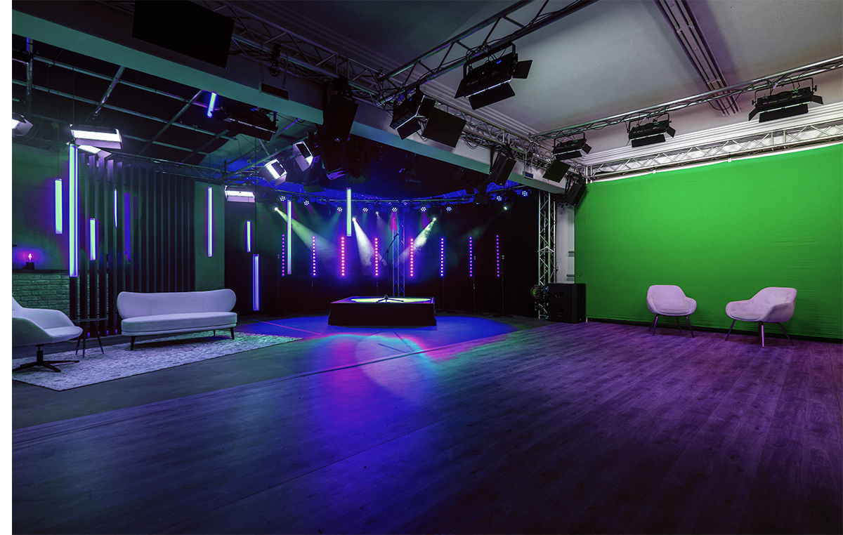 Modernes Fernsehstudio mit Bühne, Bühnenbeleuchtung und grüner Greenscreen-Wand, umgeben von Sesseln und Sofas.