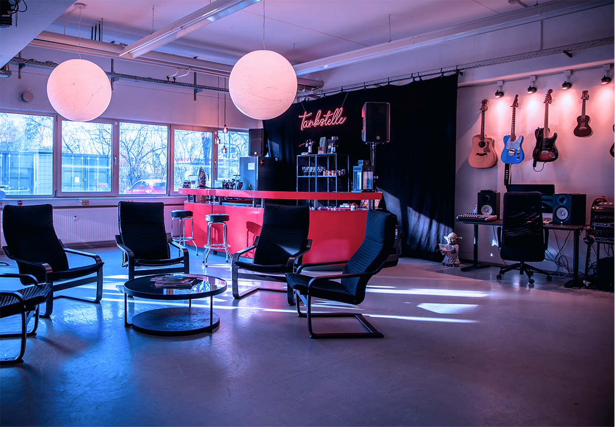 Modernes Studio mit Sitzbereich, roten Barhockern, umlaufender Bar und Wand mit sechs Gitarren bei gedämpftem bläulichem Licht.