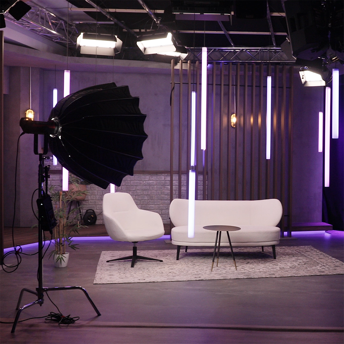 Aufnahmestudio mit einem weißen Sofa, einem weißen Sessel, einem kleinen runden Tisch und mehreren hängenden violetten und weißen LED-Röhrenlampen.