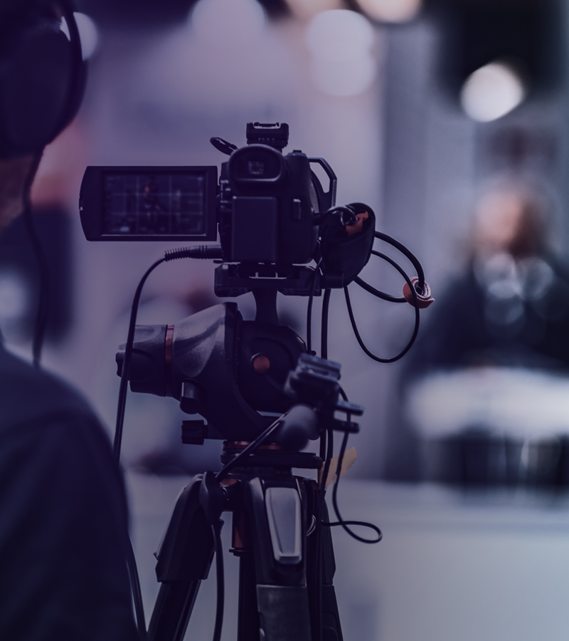 Professionelle Videokamera auf Stativ, im Hintergrund unscharfe Person mit Mikrofon.