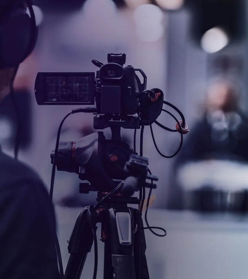 Professionelle Videokamera auf Stativ, im Hintergrund unscharfe Person mit Mikrofon.