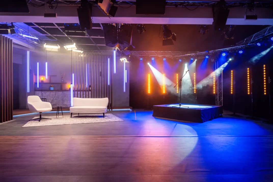 Moderne Fernsehstudio-Bühne mit weißem Sofa, Stuhl, Teppich und beleuchtetem Mikrofon auf Podium.