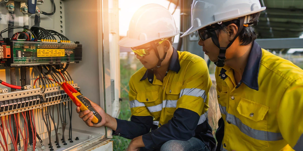 Electrical technicians in safety gear inspect industrial control panel with multimeter