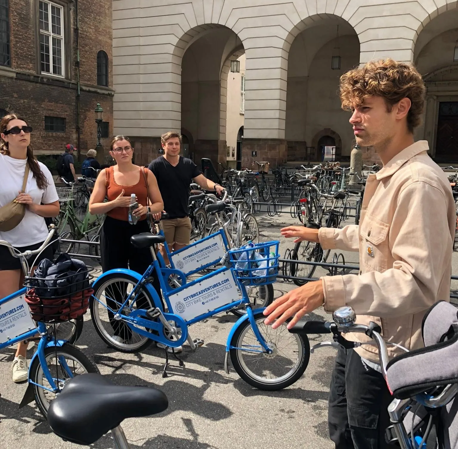 Young man in beige jacket explaining to a group of three people standing near blue city rental bikes in an urban bike parking area.