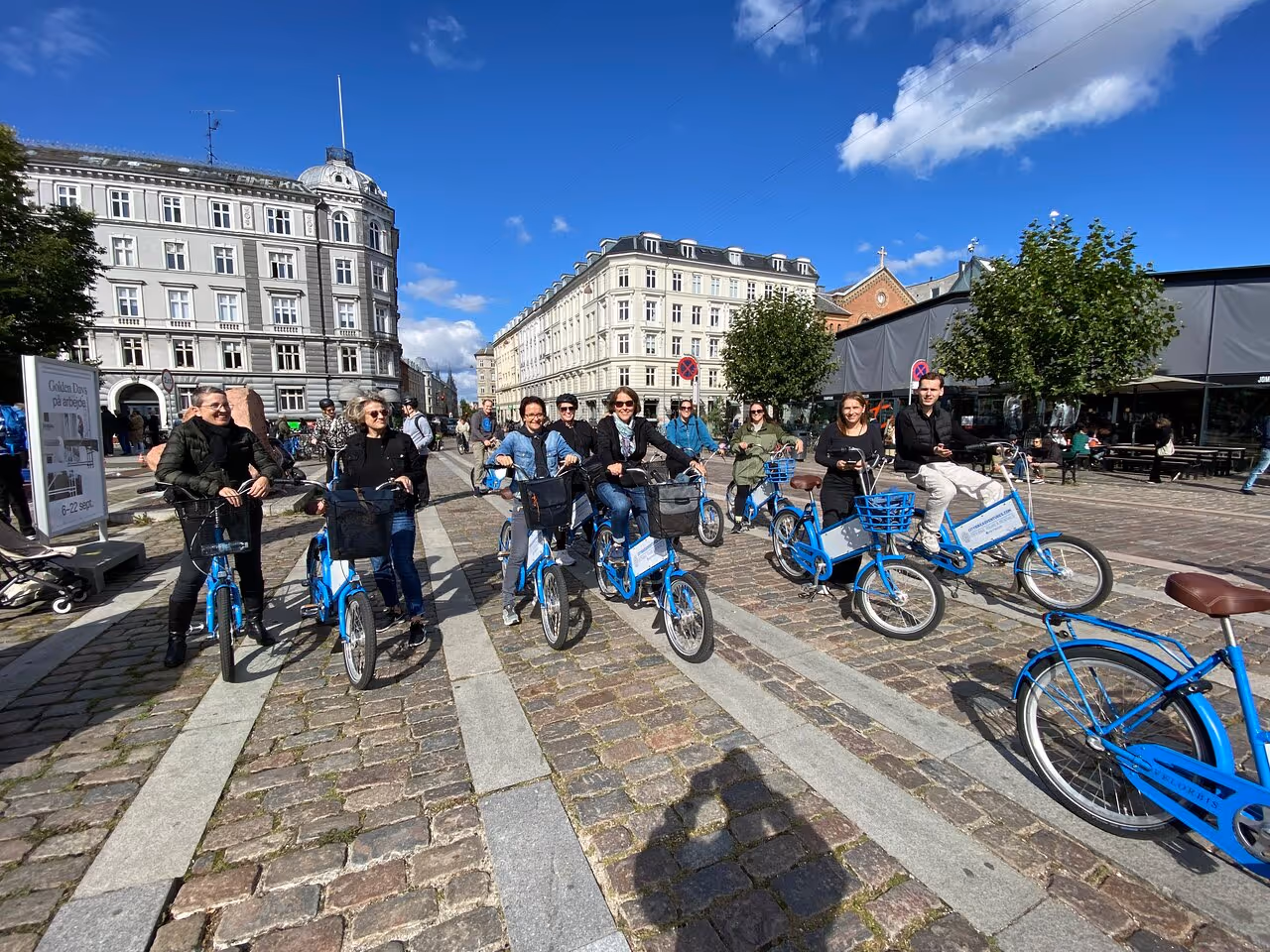 Group of people riding blue rental bikes on a cobblestone street in a European city under a clear blue sky.