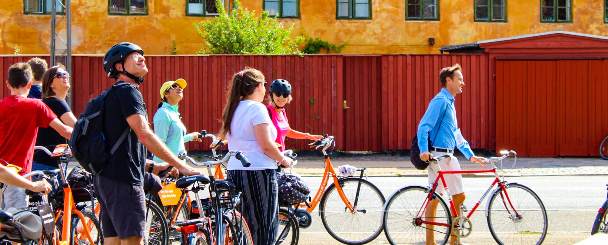 Group of people with bicycles standing on a street in front of a red fence and yellow building.