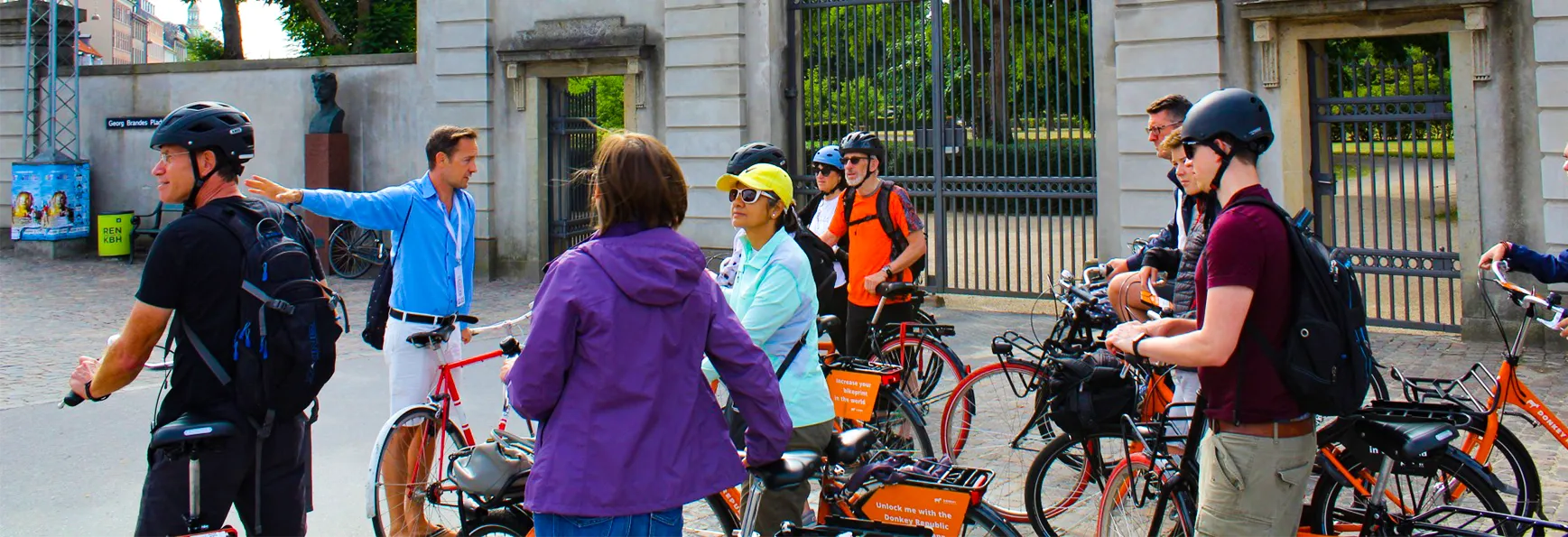 Group of cyclists wearing helmets gathered near a gated entrance, with one man in a blue shirt gesturing and talking.