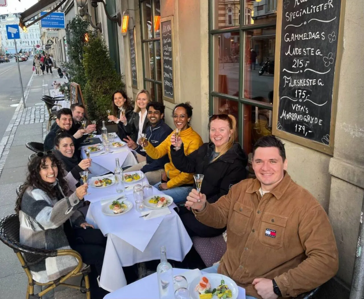 Group of nine people sitting at outdoor restaurant tables smiling and raising glasses in a toast.