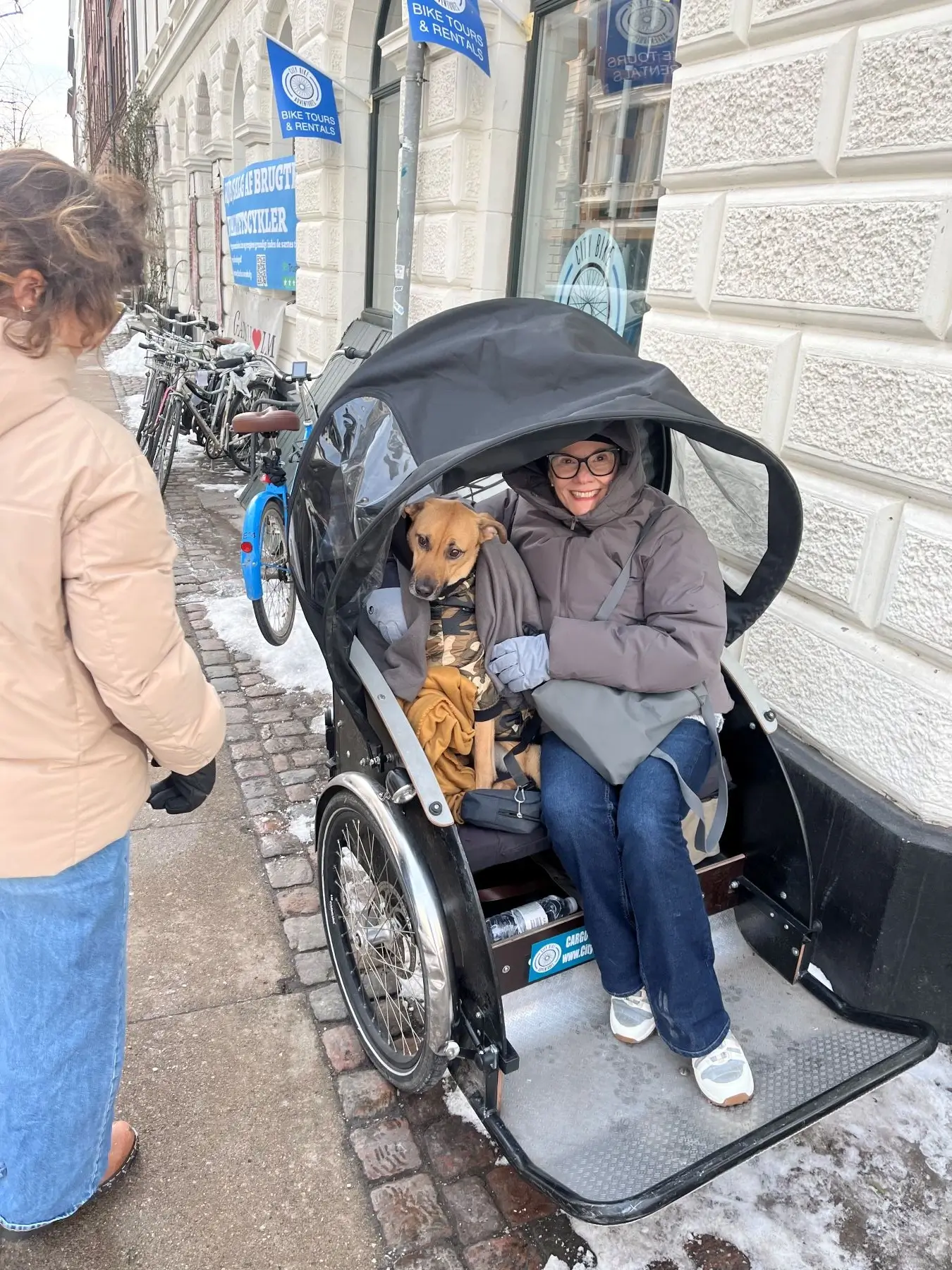 Woman wearing glasses and a gray jacket sitting in a black bike rickshaw with a dog wrapped in a blanket.