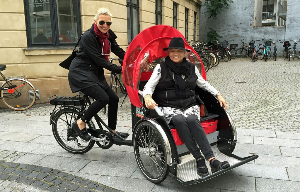 Woman riding a black trishaw with another woman seated under a red canopy in a cobblestone urban area.