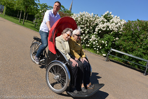 Man riding a tricycle bicycle carrying two elderly women seated in front with a red canopy in a park.