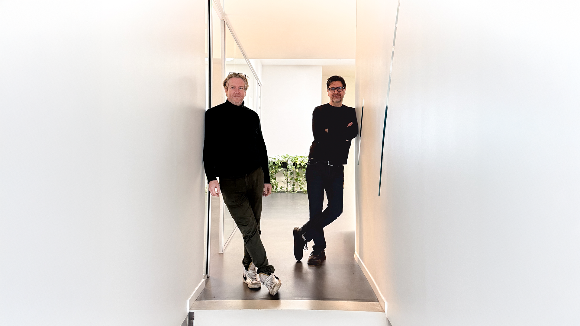 Twee mannen in donkere kledij staan ontspannen in een smalle, lichte gang met witte muren; één leunt tegen de muur vooraan, de ander staat met gekruiste armen iets verderop, met een plant zichtbaar op de achtergrond.