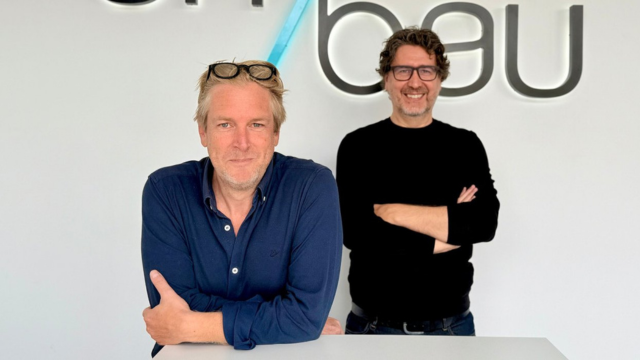 Twee mannen poseren voor een witte muur met het Umbau-logo; de man vooraan leunt op een balie in een blauw hemd met bril op het hoofd, de andere staat erachter met gekruiste armen en een zwarte trui.