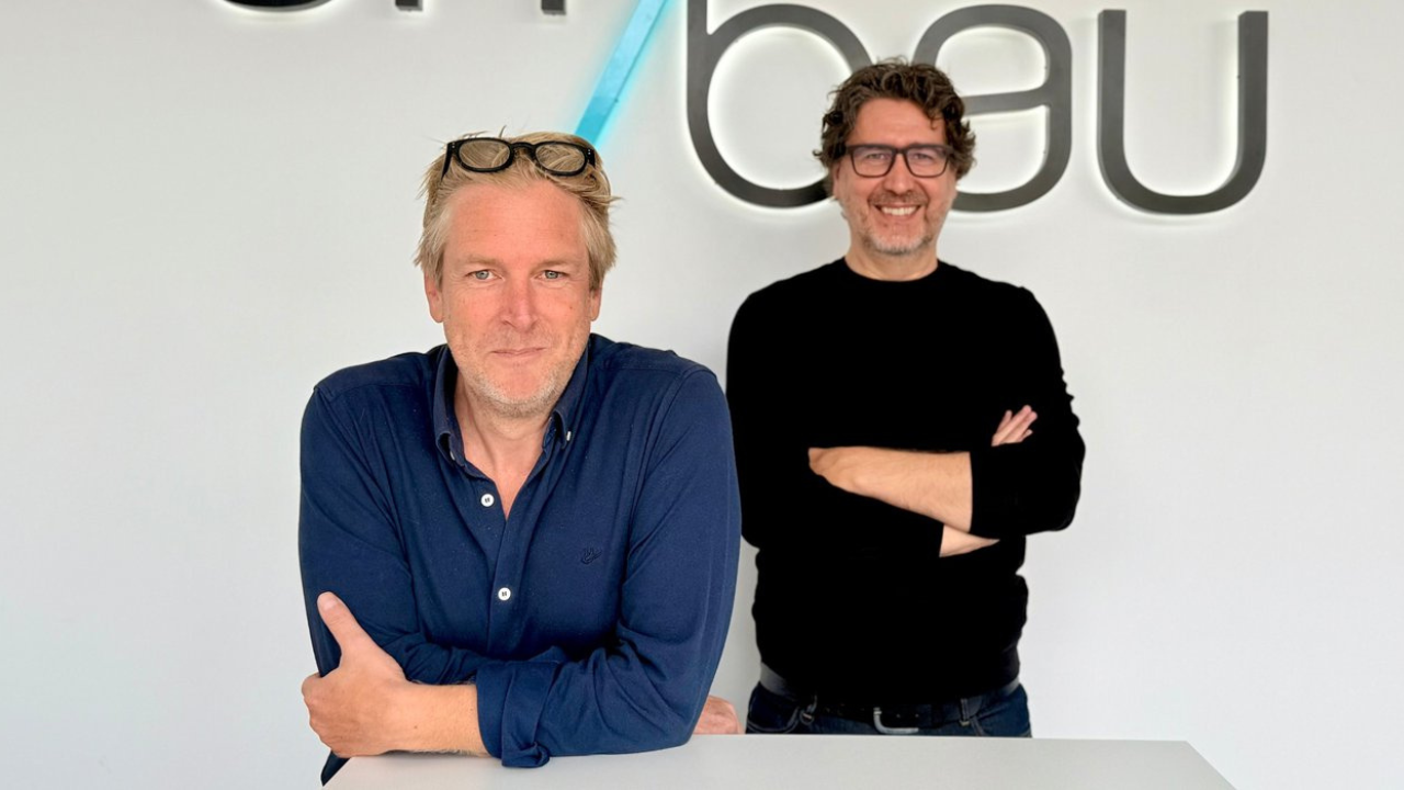 Twee mannen poseren voor een witte muur met het Umbau-logo; de man vooraan leunt op een balie in een blauw hemd met bril op het hoofd, de andere staat erachter met gekruiste armen en een zwarte trui.