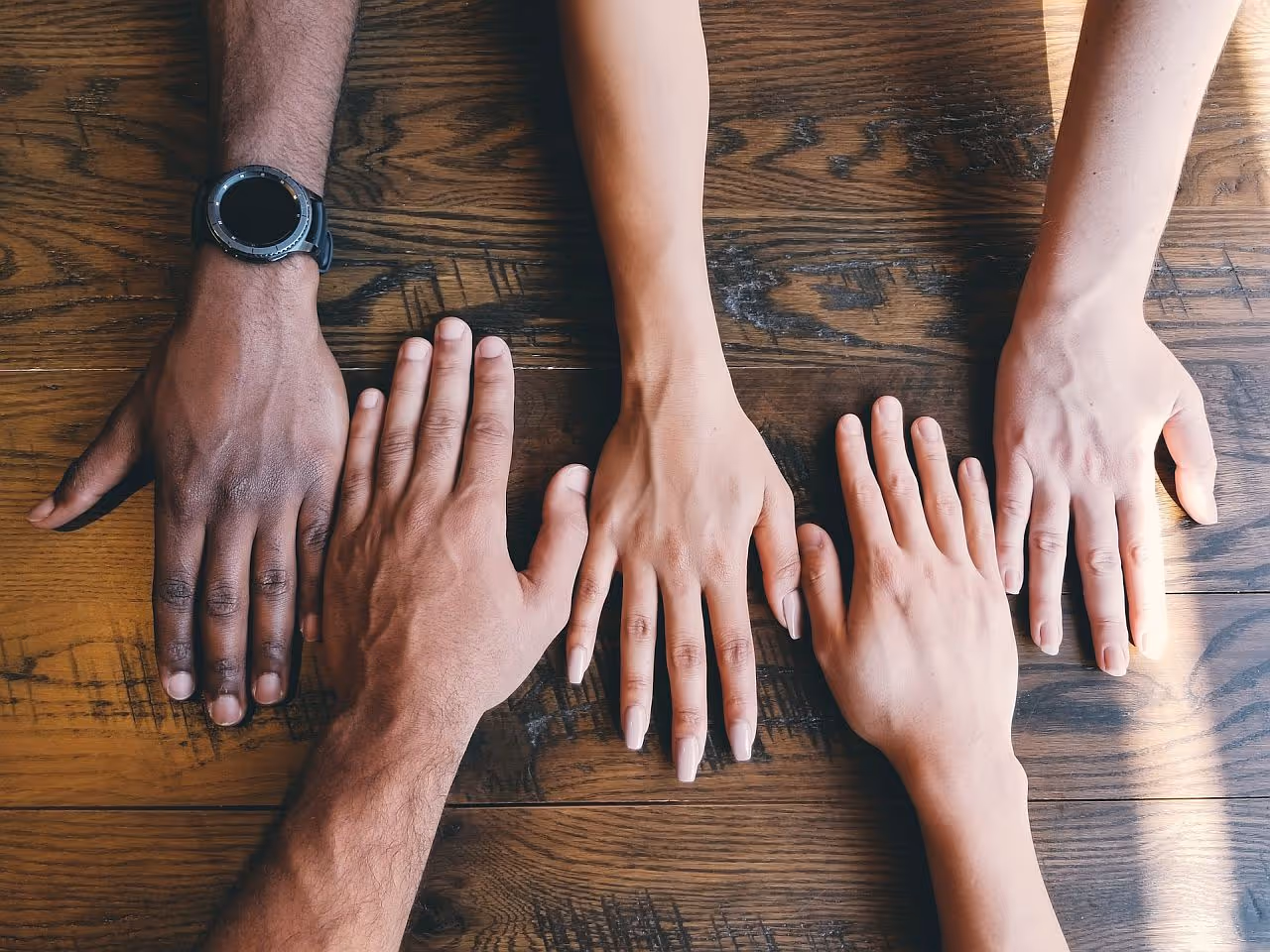 Close-up of people's hands on table; employee rewards dei concept|Two men looking over shoulder of coworker on laptop|Two women speaking to each other across small table