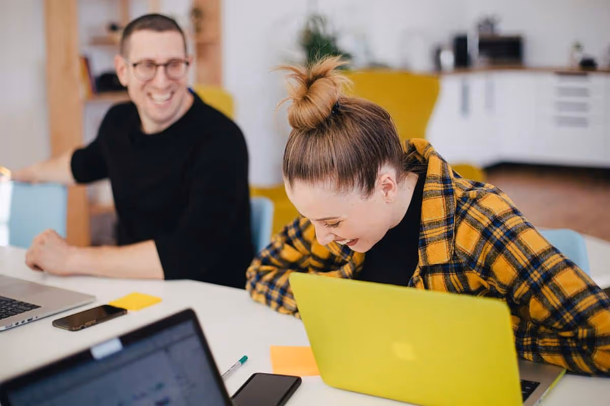 Two work colleagues laughing in the office; talent war concept|Three women talking to each other around conference table|Two women talking to each other behind conference table
