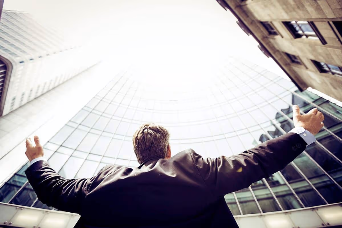 Business man lifting his arms outside, surrounded by large buildings; employee recognition concept|Young employees seated around wooden table, looking at devices and holding coffee|Man smiling in modern office kitchen