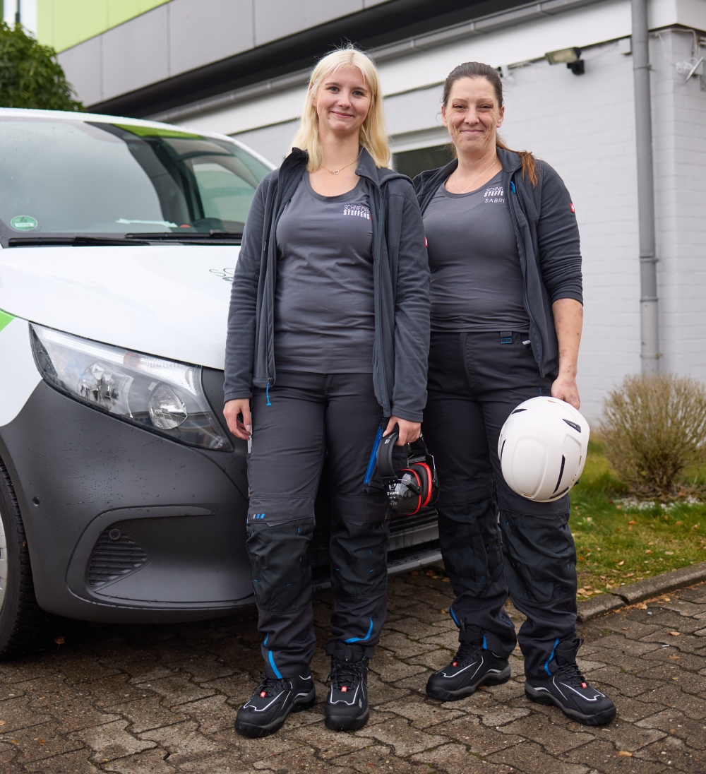 Zwei Handwerkerinnen mit Helm und Ohrenschutz vor Firmenwagen